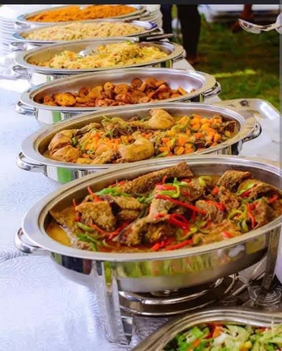 A long outdoor buffet line of silver chafing dishes filled with rice, proteins and vegetables