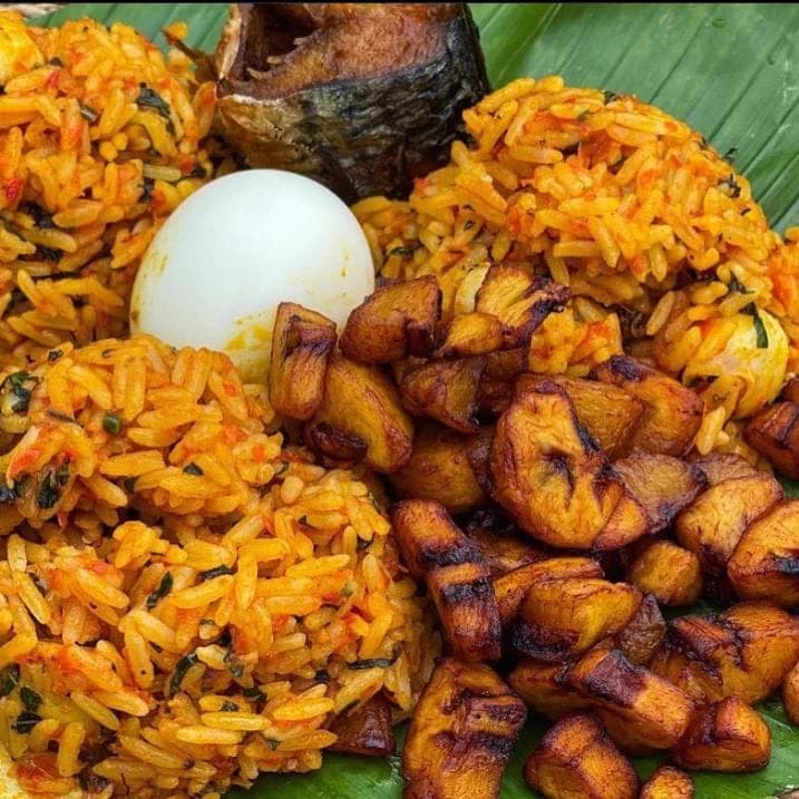 Jollof rice served on a banana leaf with fried plantain, fried fish and boiled egg