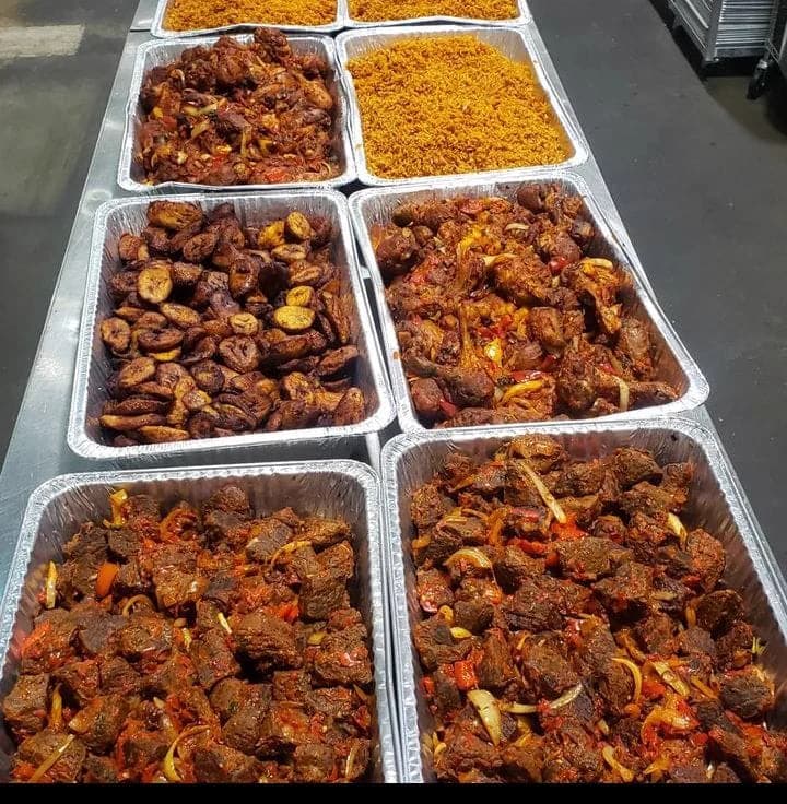 A prep table lined with food trays of jollof rice, fried plantain and peppered beef